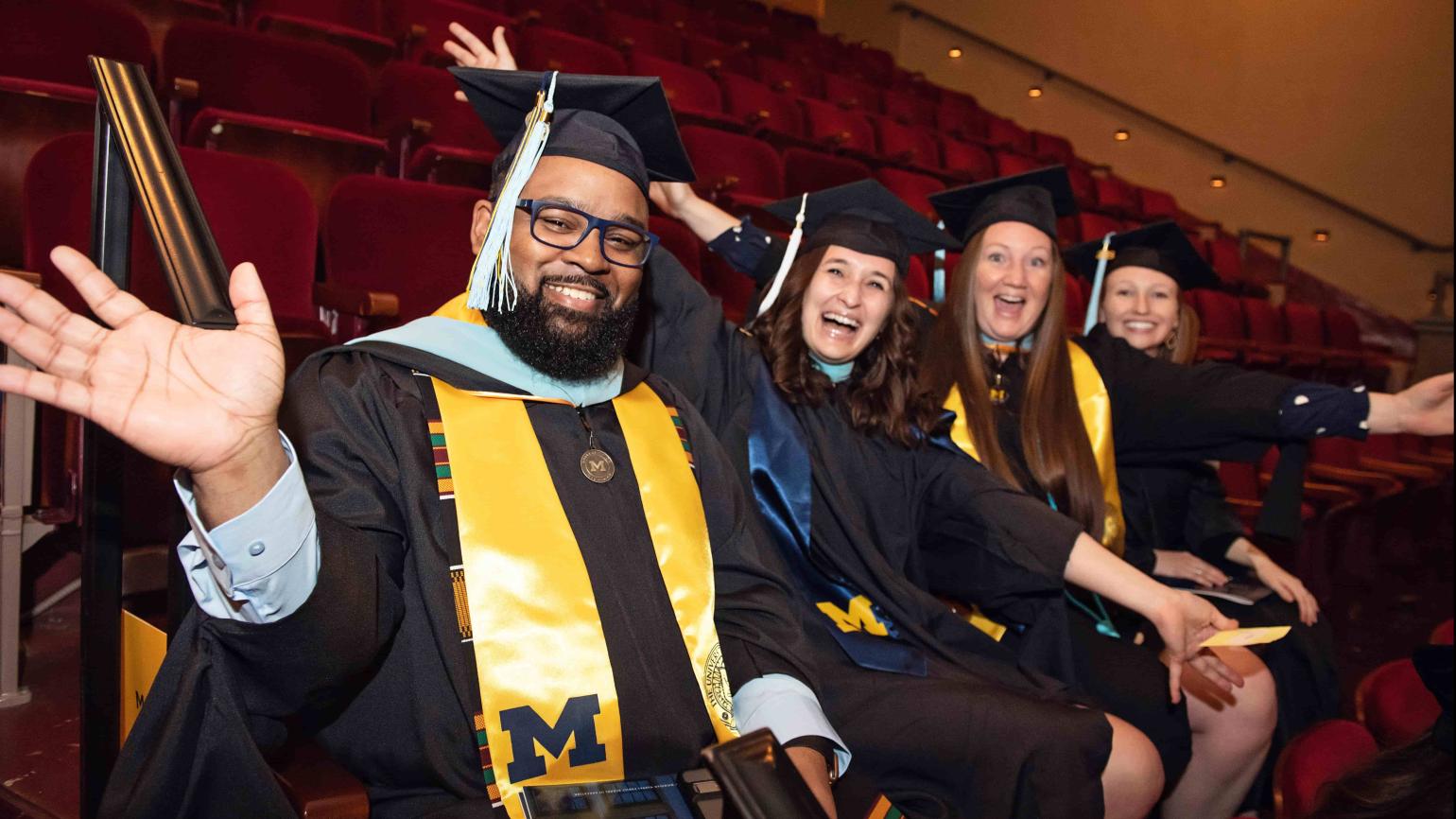 Four graduates sitting in red auditorium-style seats wave and cheer excitedly for a photo.