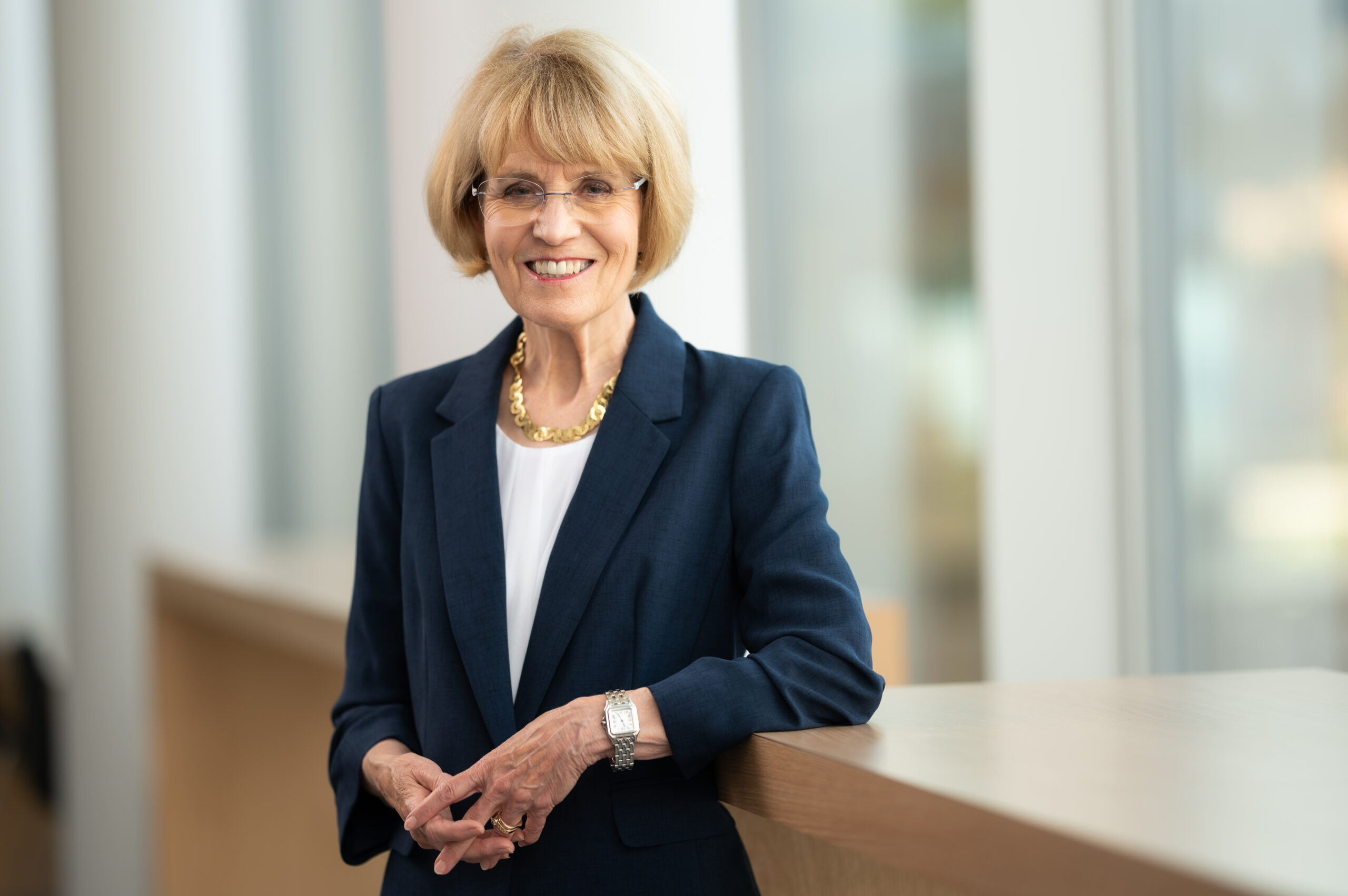 Mary Sue Coleman, wearing glasses, a white blouse, a navy blue jacket, and gold necklace, smiles and poses indoors, with one arm on a banister and her hands clasped in front of her, for a photograph.