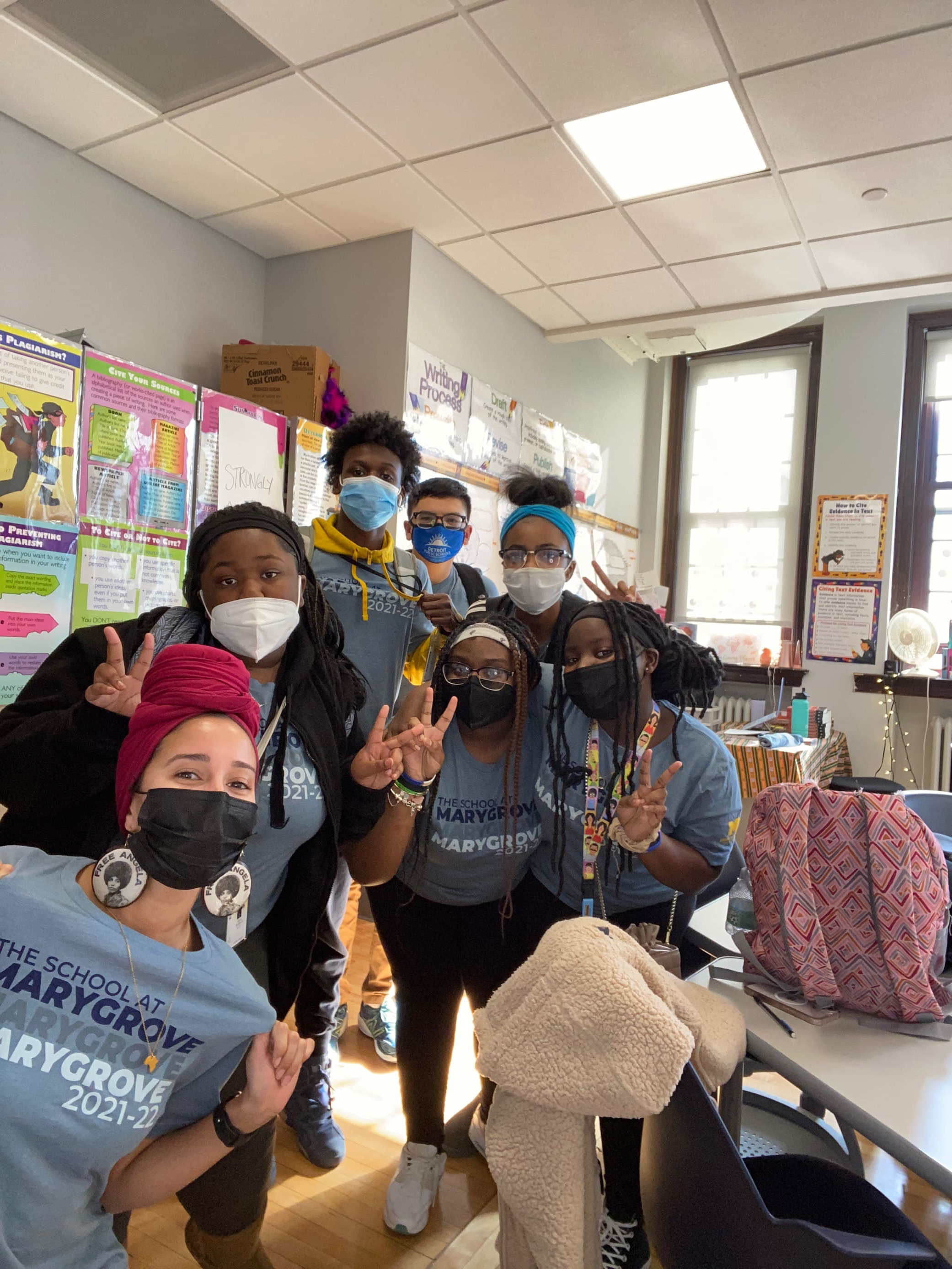 Several masked students, faculty, and staff, wearing Marygrove shirts and throwing peace signs, pose for a photo in a classroom.
