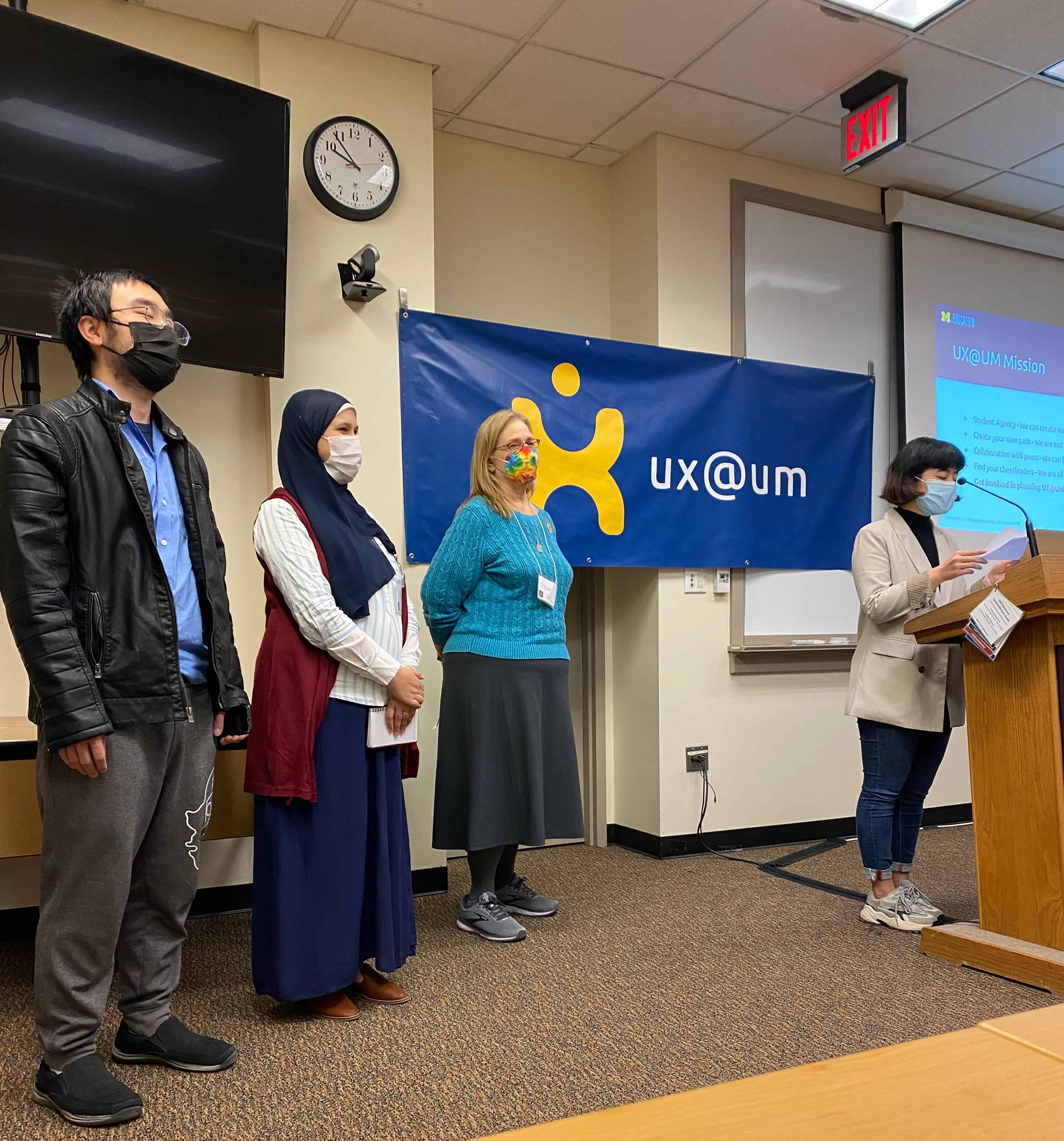 Presenters, wearing masks, standing at a podium in the front of a classroom.