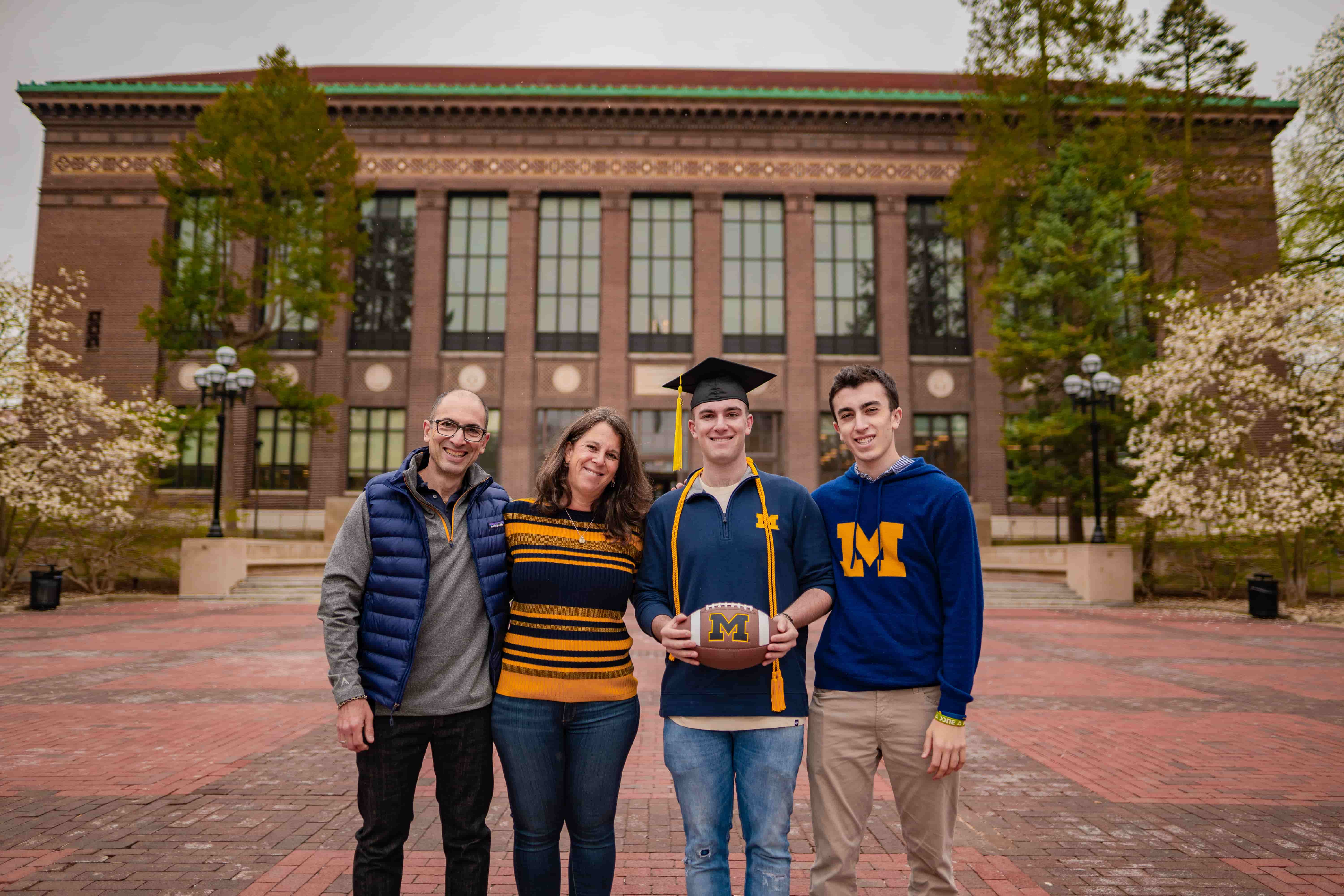 Graduate wearing cap and holding a football smiles in front of Hatcher Graduate Library with three family members.
