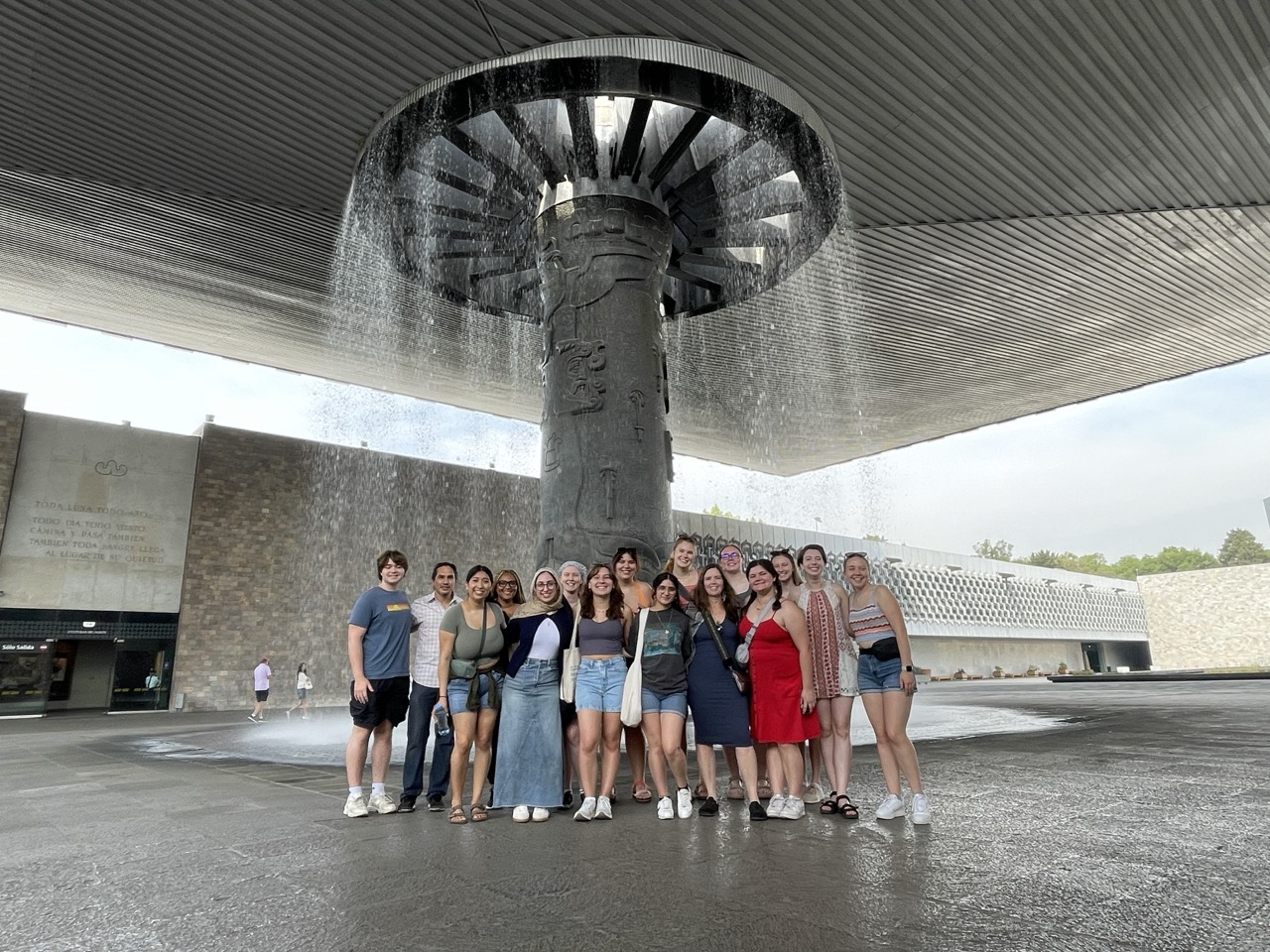 The whole group of ELD interns stand in front of a fountain at the Mexican Museum of Anthropology.  Water falls behind the group as they huddle with a smile.