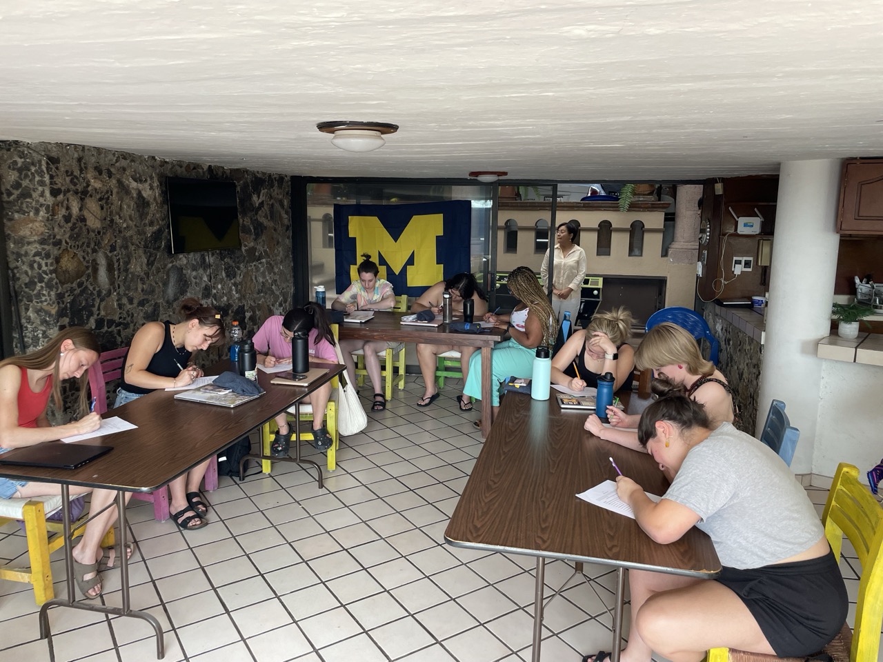 Interns seated at tables in the ELD abroad classroom, a room with a tile floor and a large block M flag.