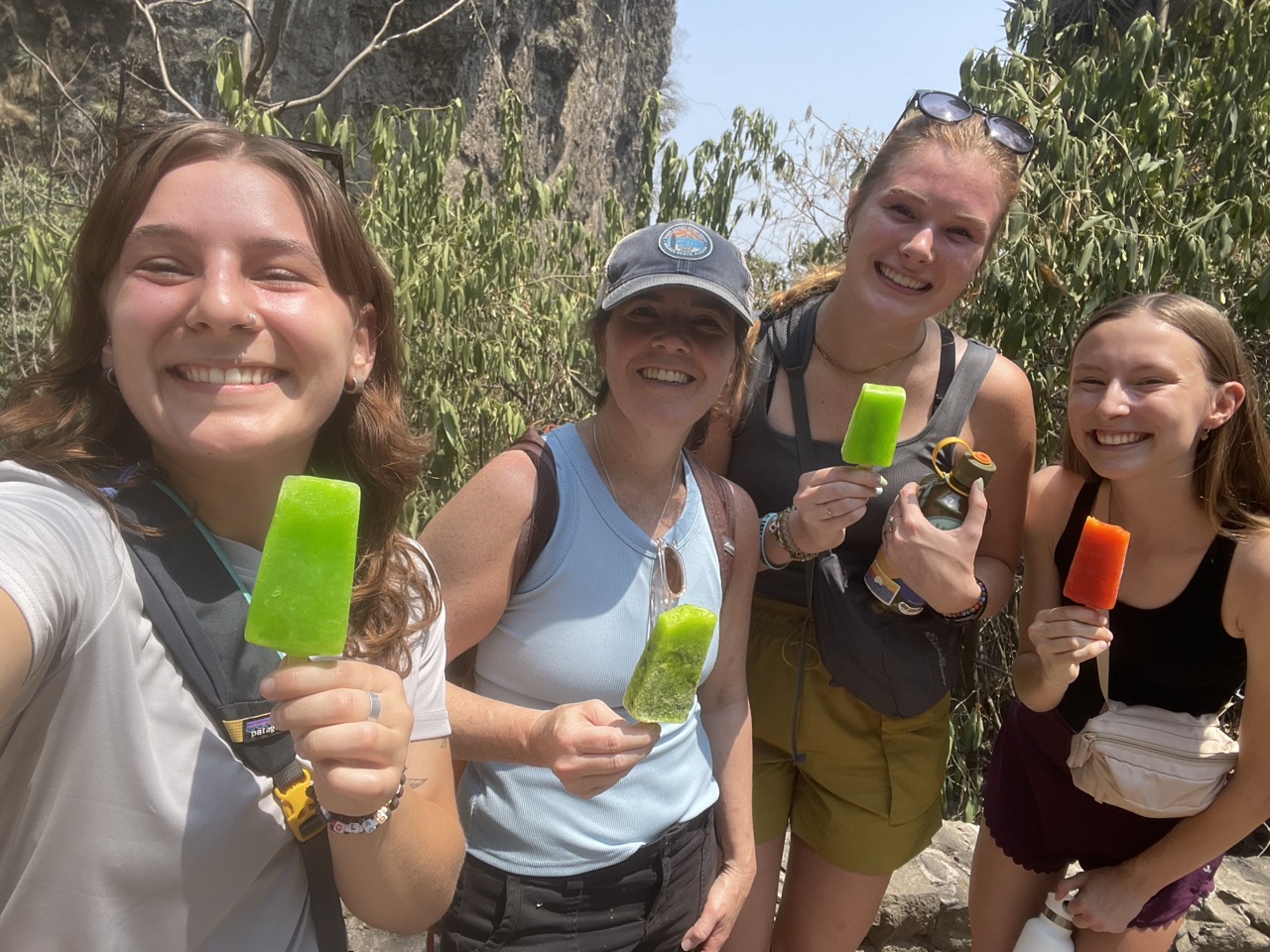 Riley, Susan, Julia and Londyn smile with popsicles in their hands during a stop on a hike in the Tepoztlan mountains. Rock and greenery is visible behind them.