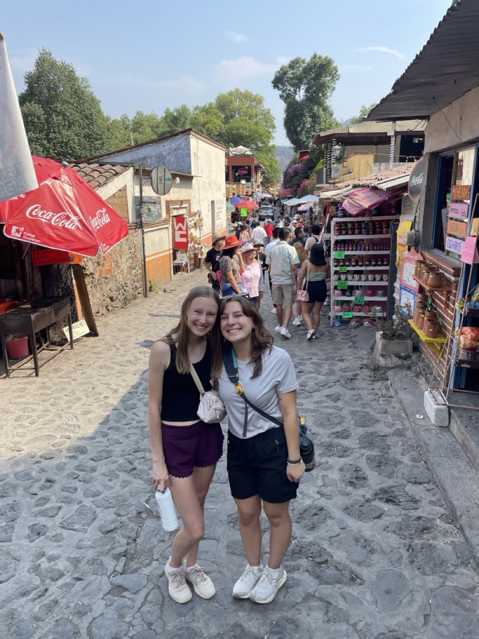 Riley and Londyn stand on the cobblestone streets of Tepoztlan, Mexico. Street vendors are visible in the background.