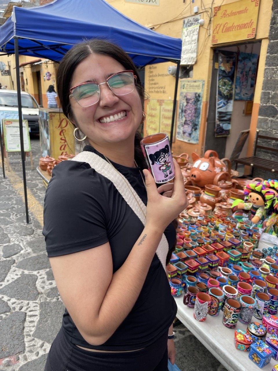 Rosie poses in front of a table of pottery souvenirs in Tepoztlan, Mexico. She smiles while holding a cup with a painted image of a girl.