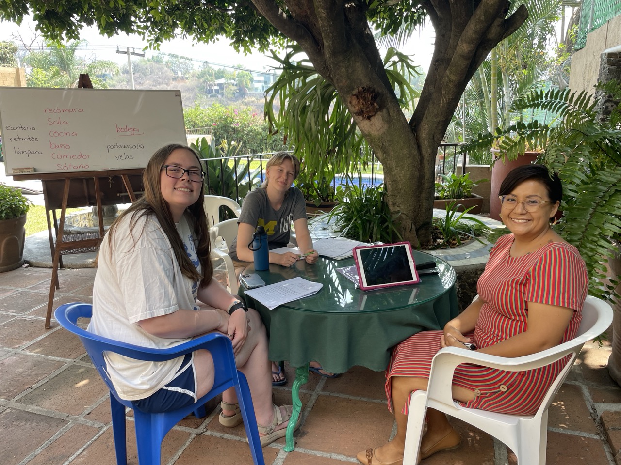 Two interns pose at an outdoor table with their Spanish teacher during a language lesson. A tree and whiteboard are visible in the background.