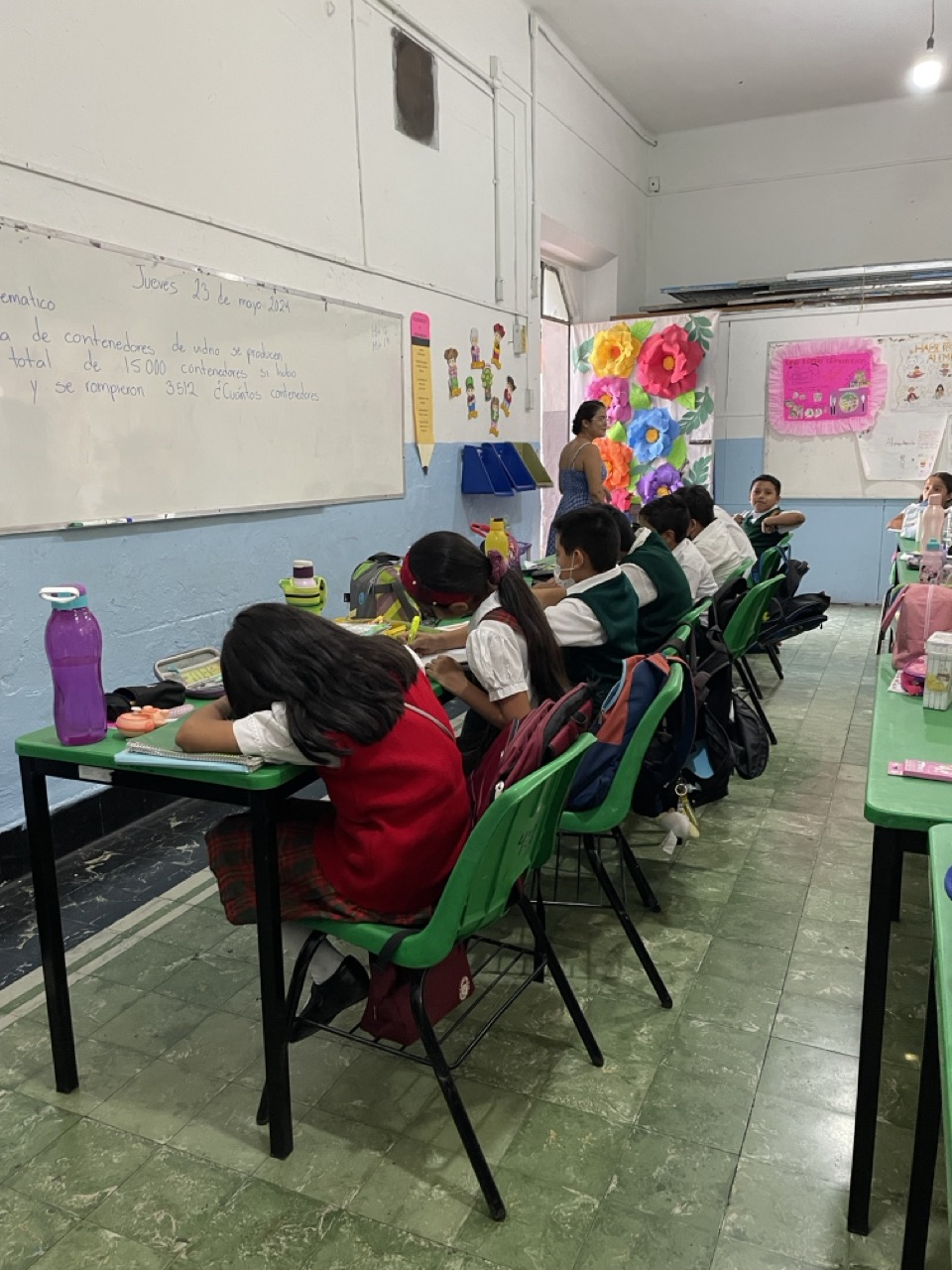 Five Mexican school children sit in a row of desks working on an assignment. Their teacher is visible in the background as she checks their work.