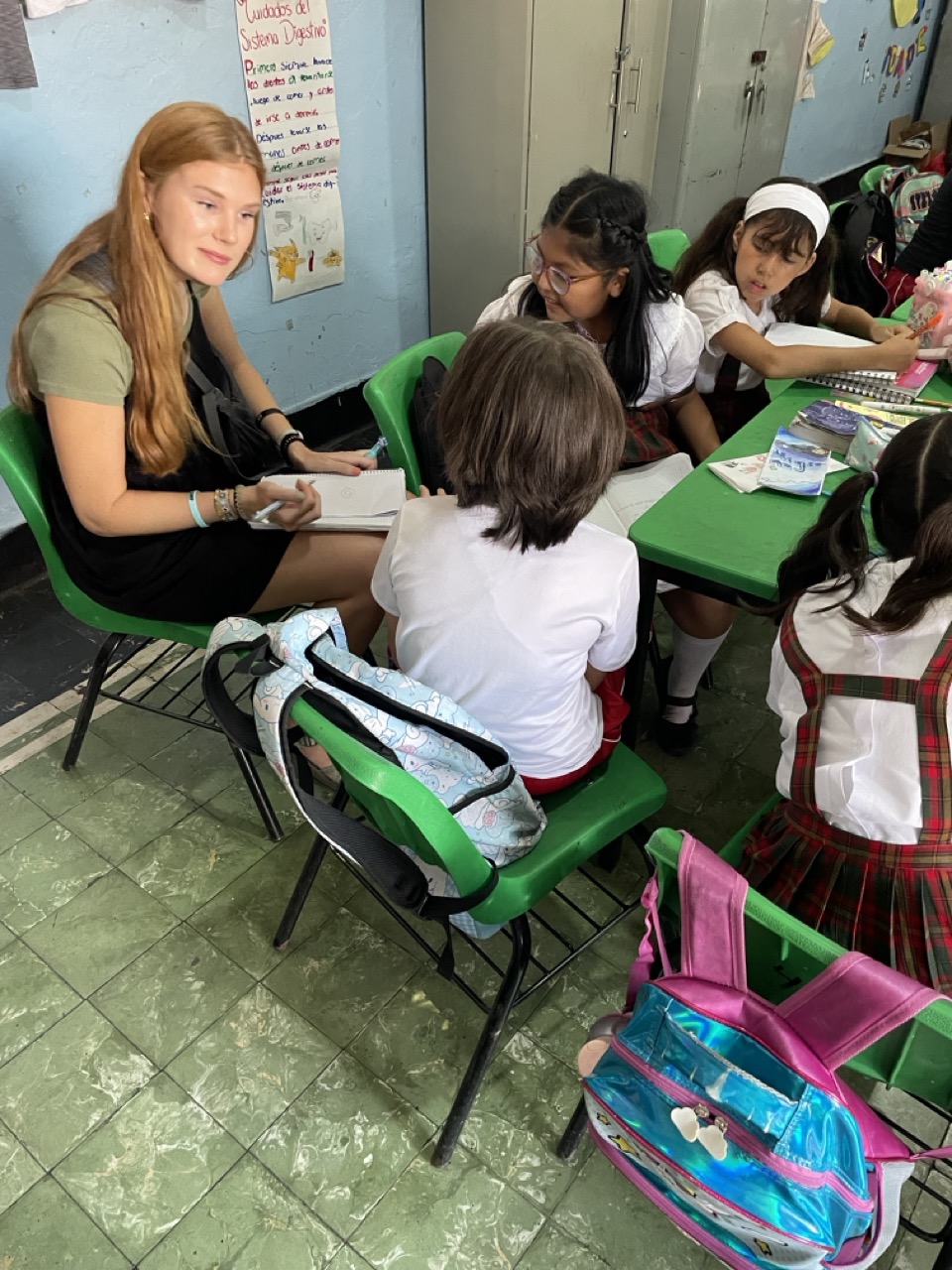 Julia is seated in a green chair next to a Mexican school girl as they engage in conversation and shared writing.