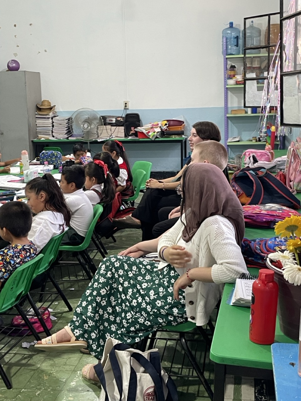 Three interns sit in the back of a Mexican school classroom and watch the teacher deliver a lesson to a group of students, seated in the row in front of them.