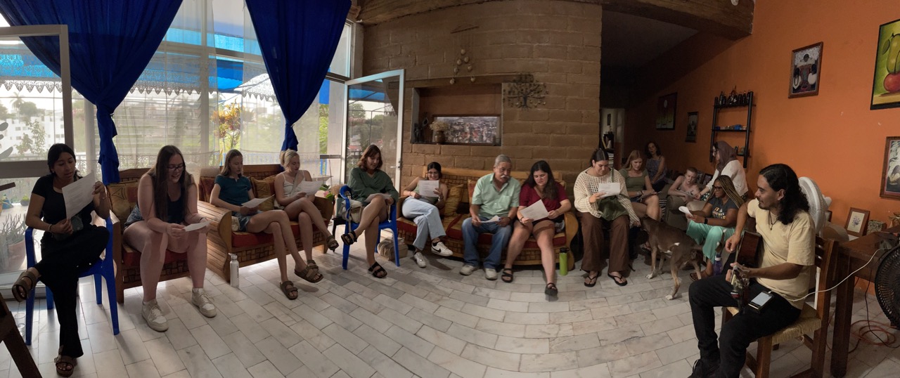 This panoramic image shows the intern group seated in a large circle, learning songs during a Mexican music class. Daniel is visible in the front, playing the guitar.