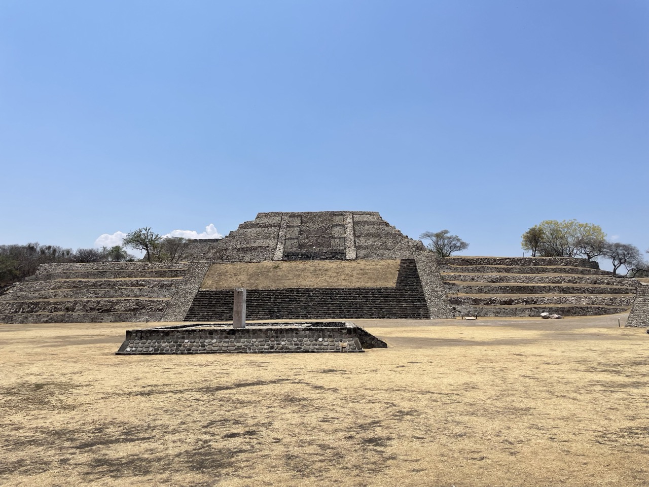 The pyramid at Xochicalco archaeological site is low to the ground, with three different sets of stairs. It is situated on the edge of a dry, grass plaza.