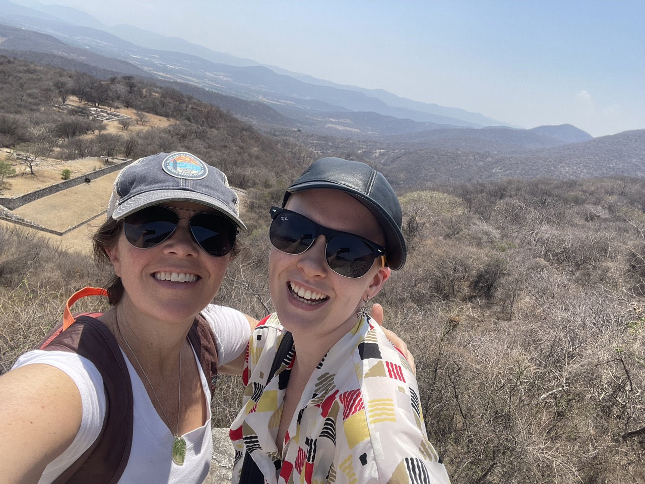 Susan and Asa smile at the top of a pyramid at Xochicalco wearing hats and sunglasses. The dry grasses and bushes of the area are visible in the background.