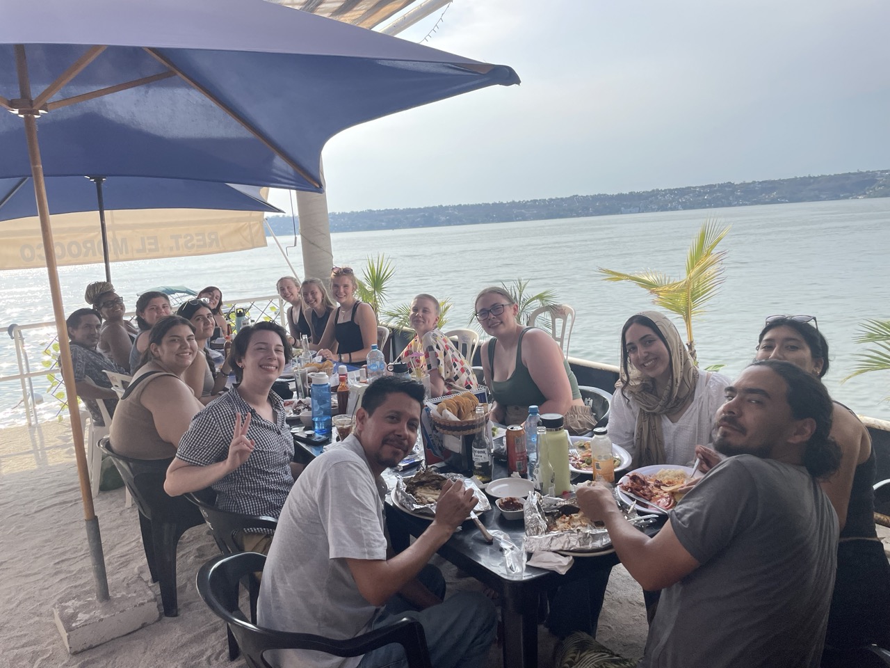 A large group of ELD interns sit at a table eating lunch in front of Lake Tequesquitengo. A blue umbrella offers shade on the visibly sunny day.