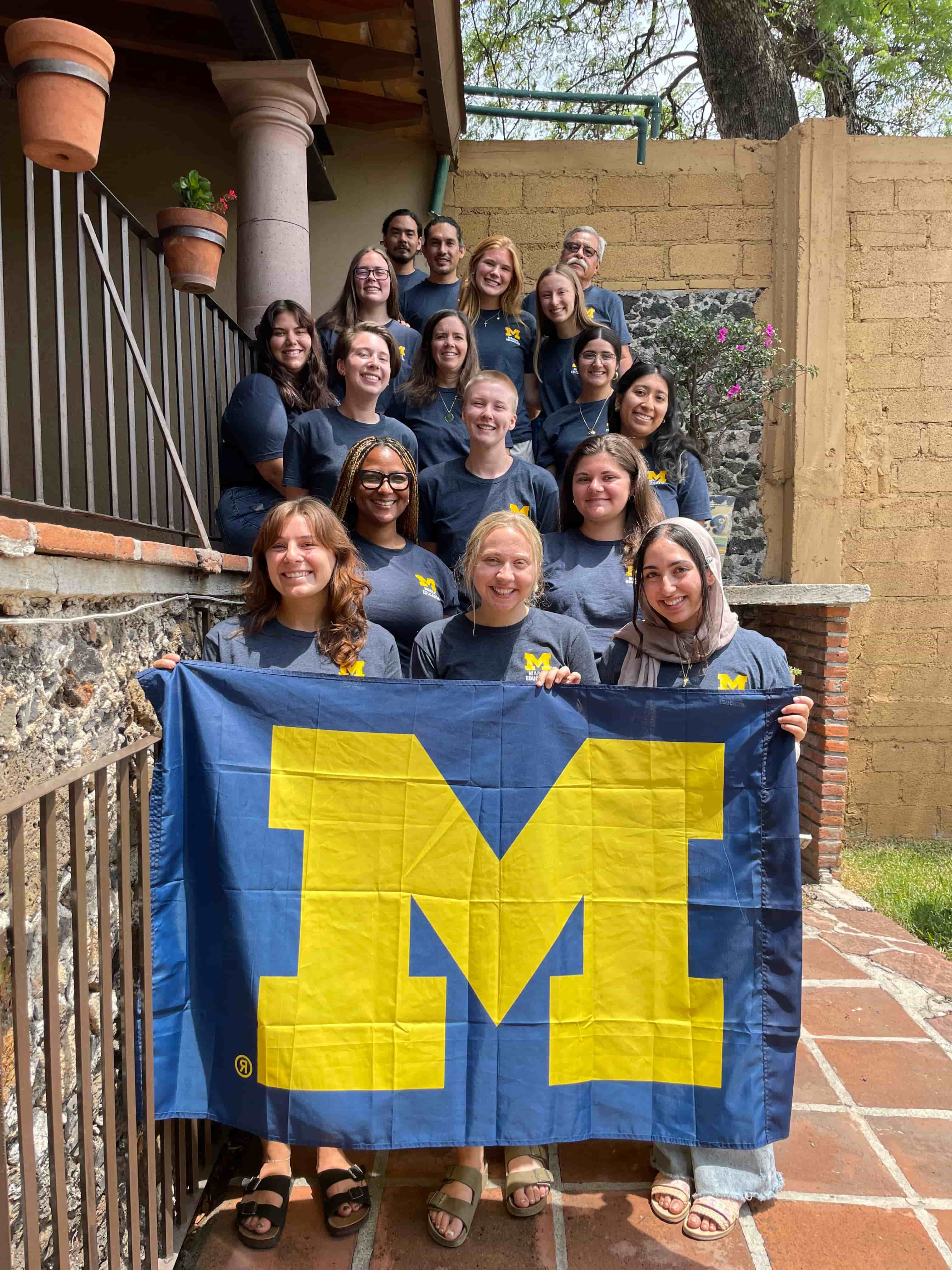 The whole ELD intern group gathers on stairs and poses in front of the block M flag on our last day of class. Everyone is wearing their U of M ELD abroad blue t-shirt.