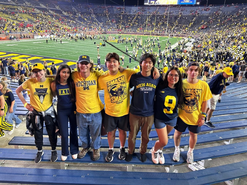 Seven LEAPS students stand with their arms around each other in the bleachers at a U-M football game