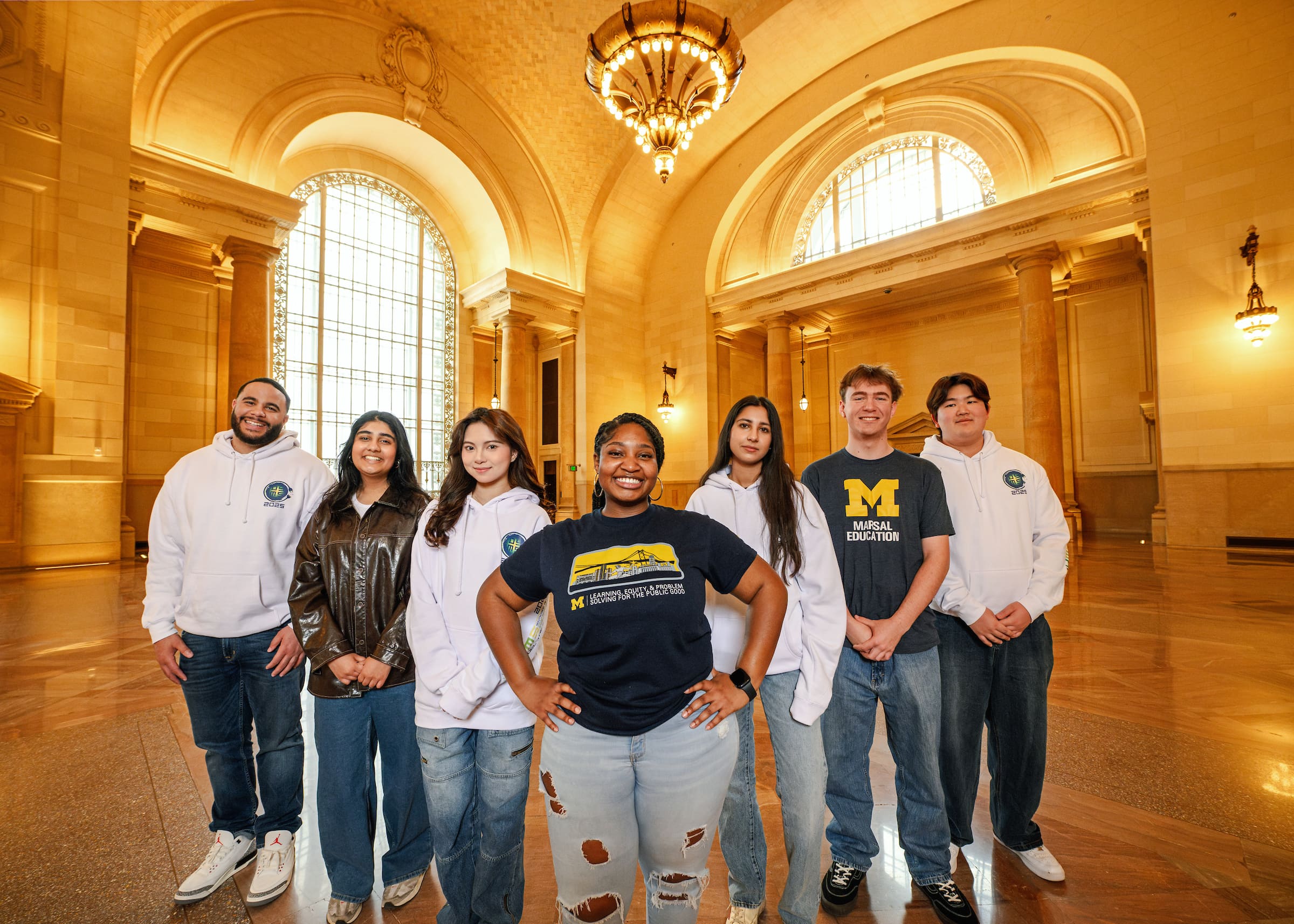 Seven people pose for a photo underneath a chandelier in a well-lit room with large windows