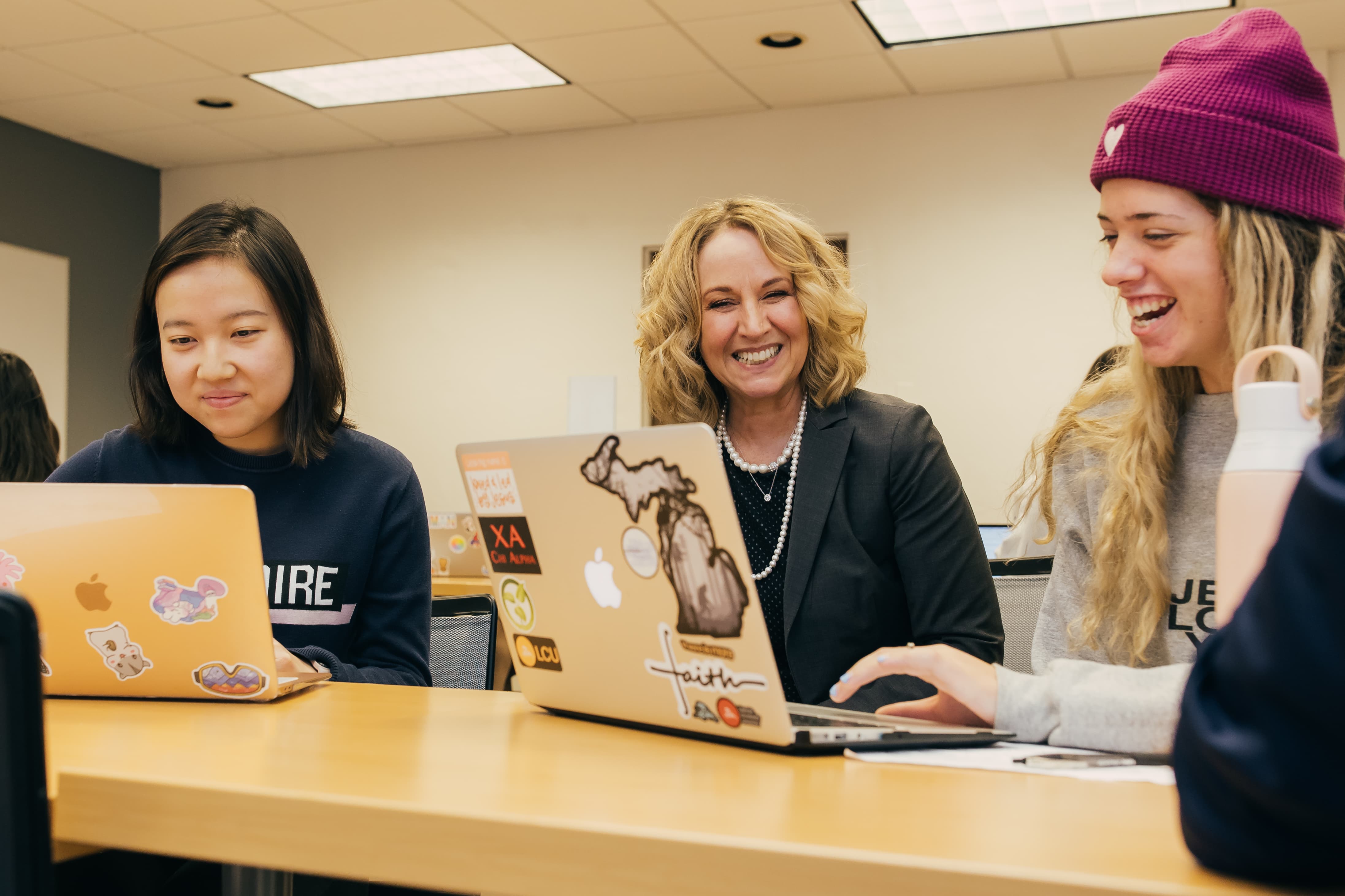 Two students sit at a desk on either side of Dean Moje. The students are working on their laptops, and the Dean is smiling broadly with them.