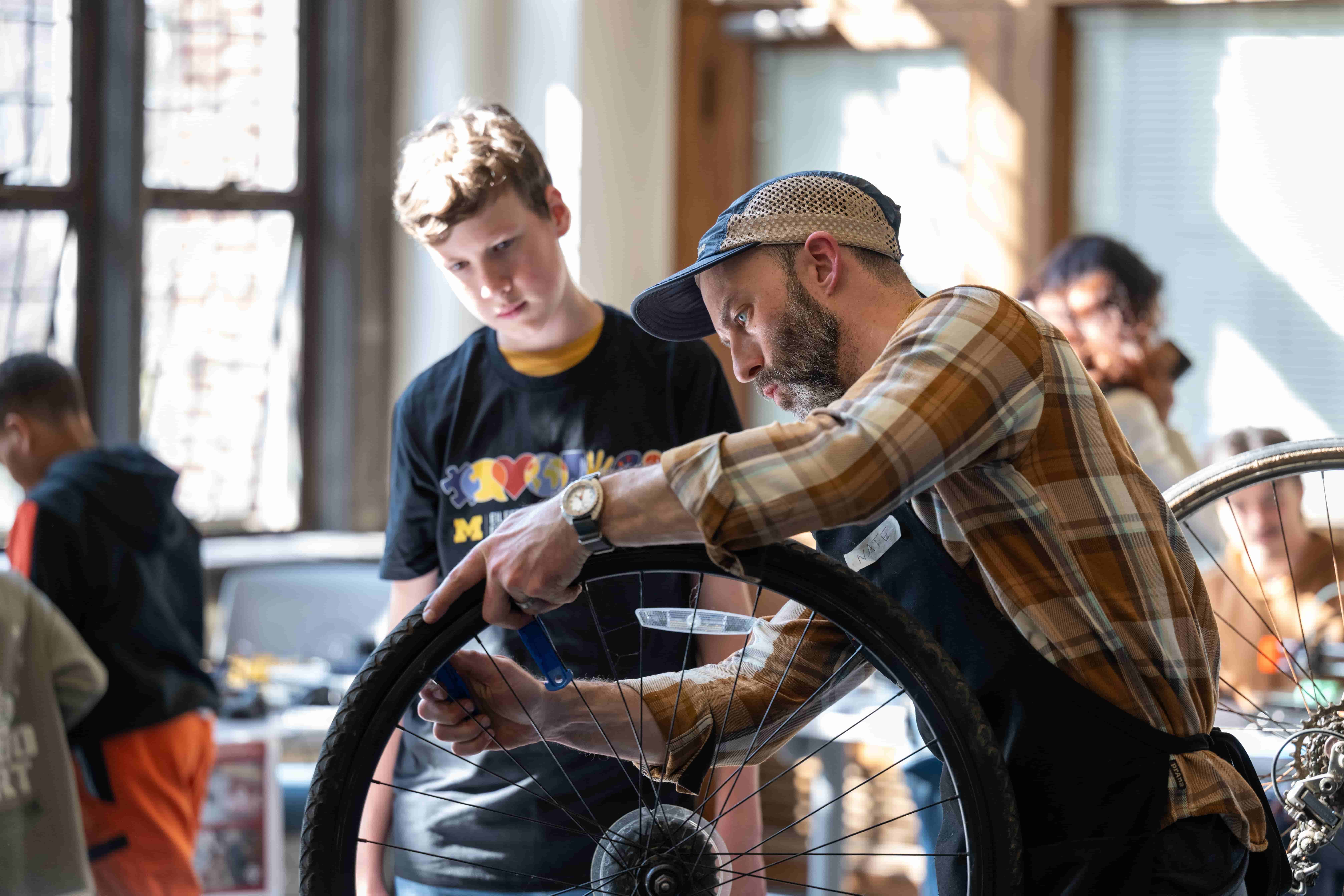 A man with a grey beard wearing a baseball cap, a brown plaid shirt, and a workshop apron appears in the foreground, adjusting the spokes on a bicycle wheel. Behind him stands a teenage boy, who watches the demonstration on bicycle maintenance. 