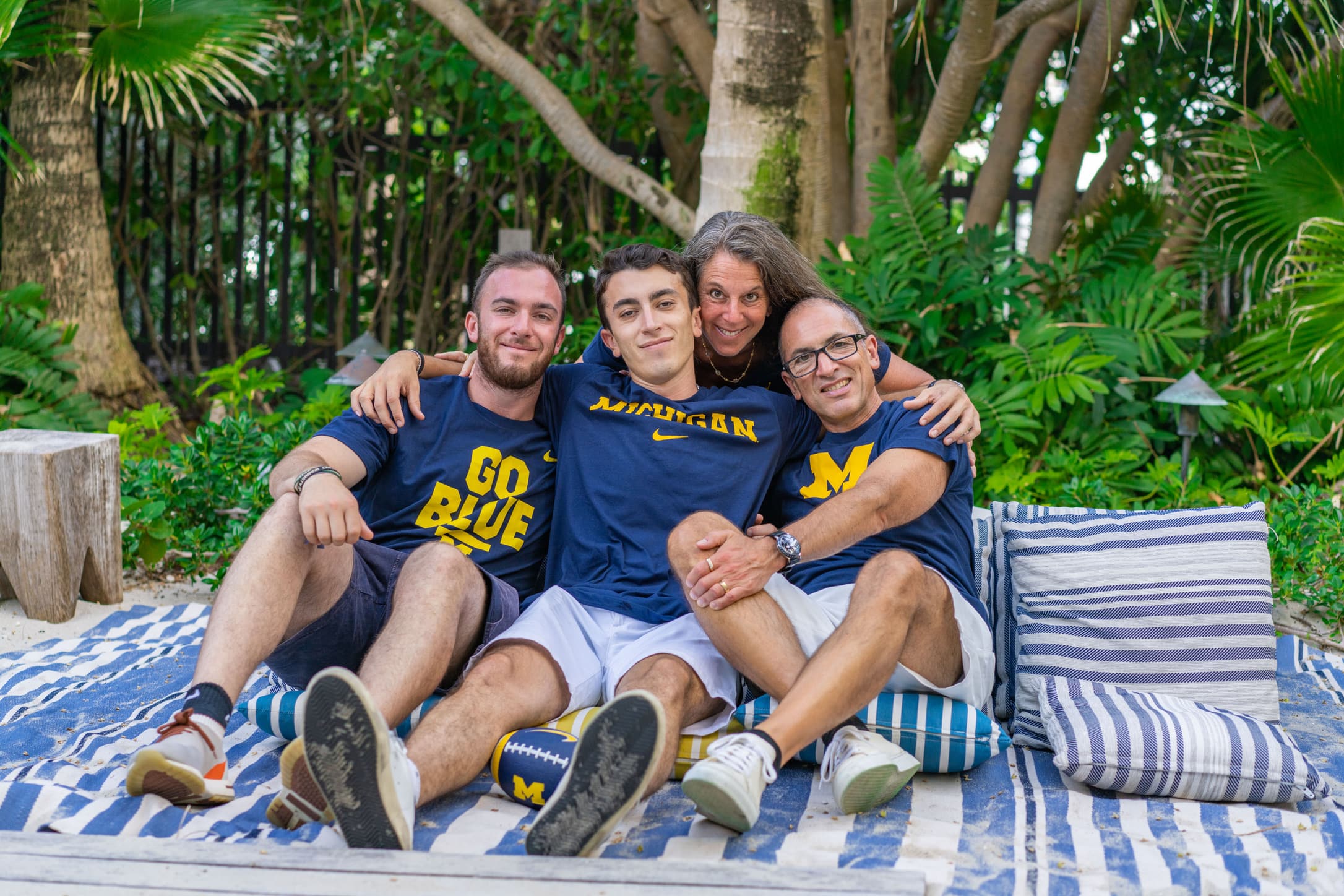 A family of four sit on a blue and white striped beach blanket in a tropical setting. Two young adult sons sit next to their mother and father. All four wear University of Michigan maize and blue apparel. 
