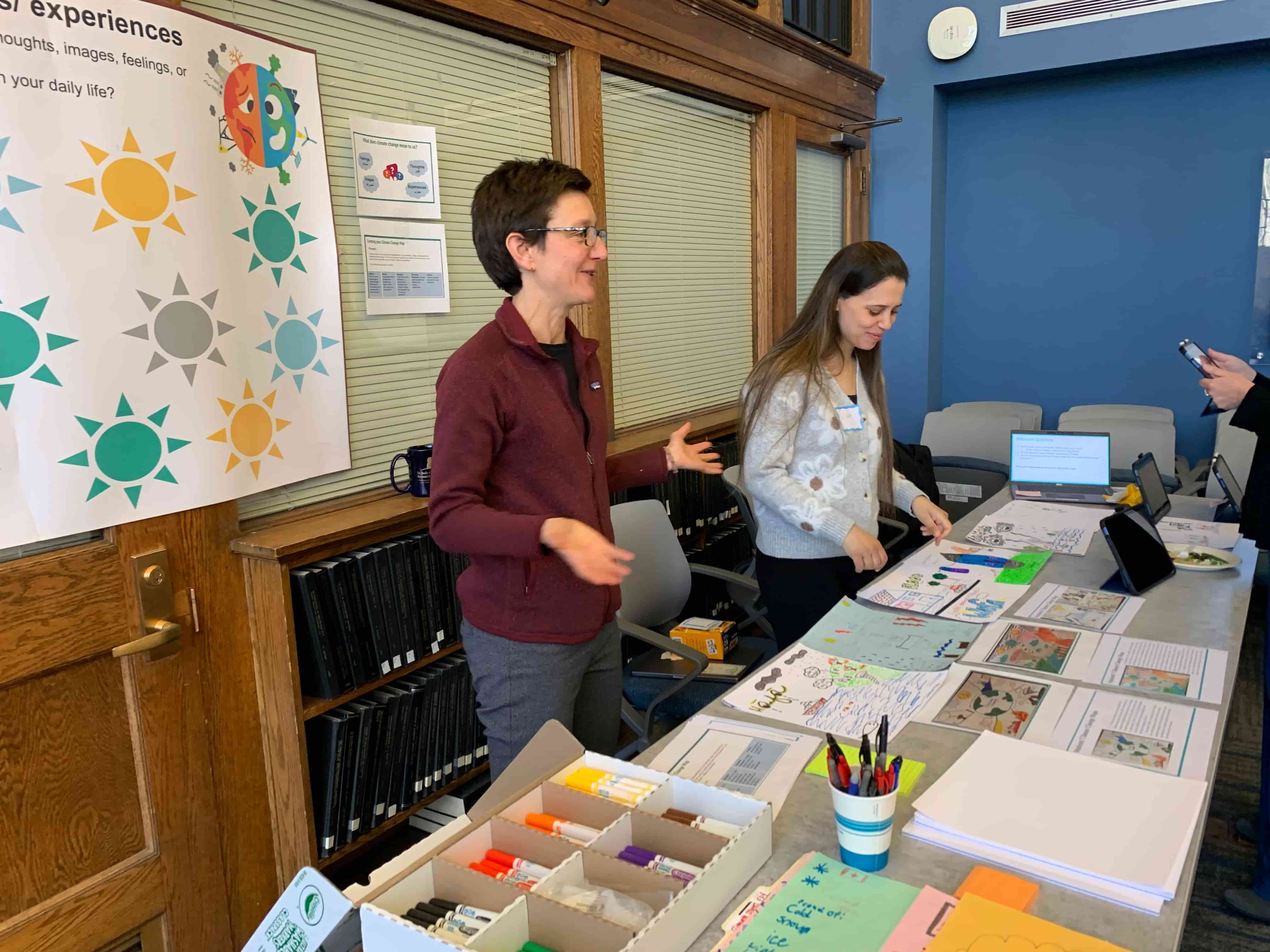 Educational Studies Chair Angela Calabrese Barton stands behind a display table with her arms and hands open, explaining the colorful drawings, worksheets, and informational papers on the table. 
