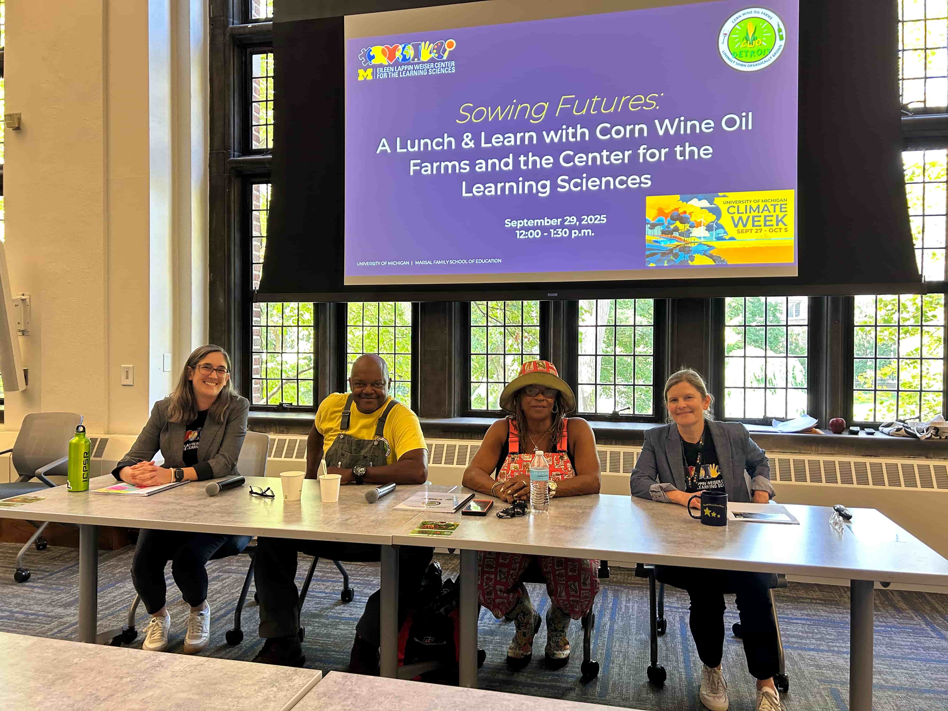 Four speakers sit at a conference table in front of a large window. Behind them, a presentation screen displays information for “Sowing Futures: A Lunch & Learn with Corn Wine Oil Farms and the Center for the Learning Sciences,” sponsored by the Eileen Lappin Weiser Center for the Learning Sciences. 