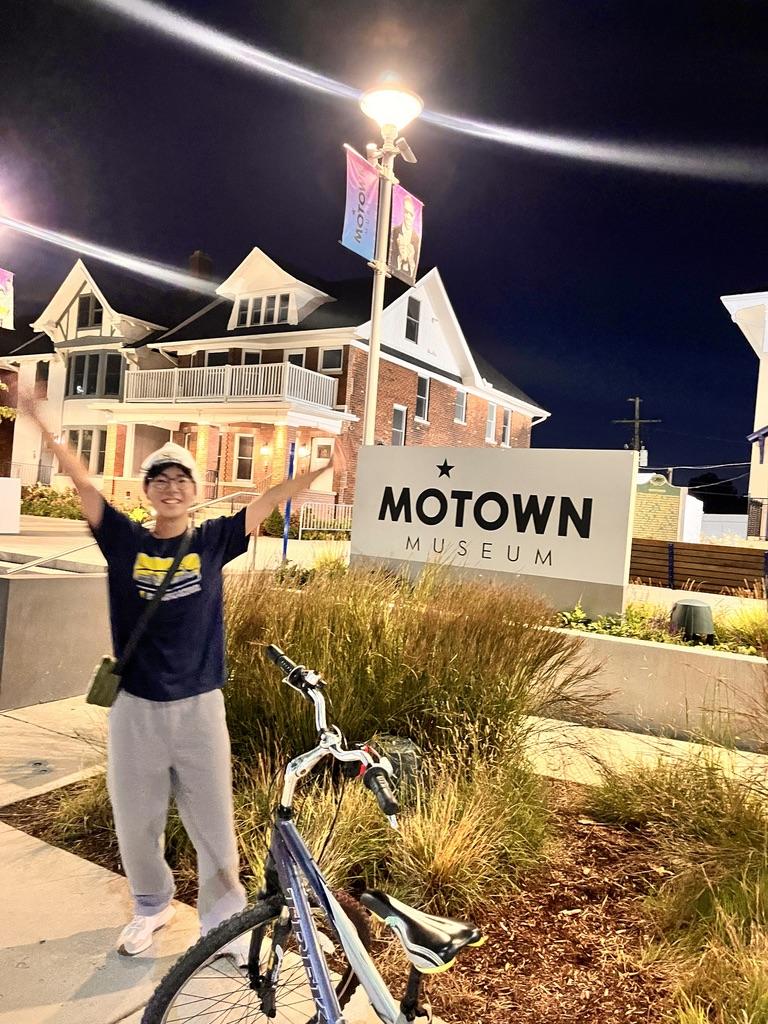 A male student stands with his arms outstretched in front of the Motown Museum sign. A bike is in the foreground. 