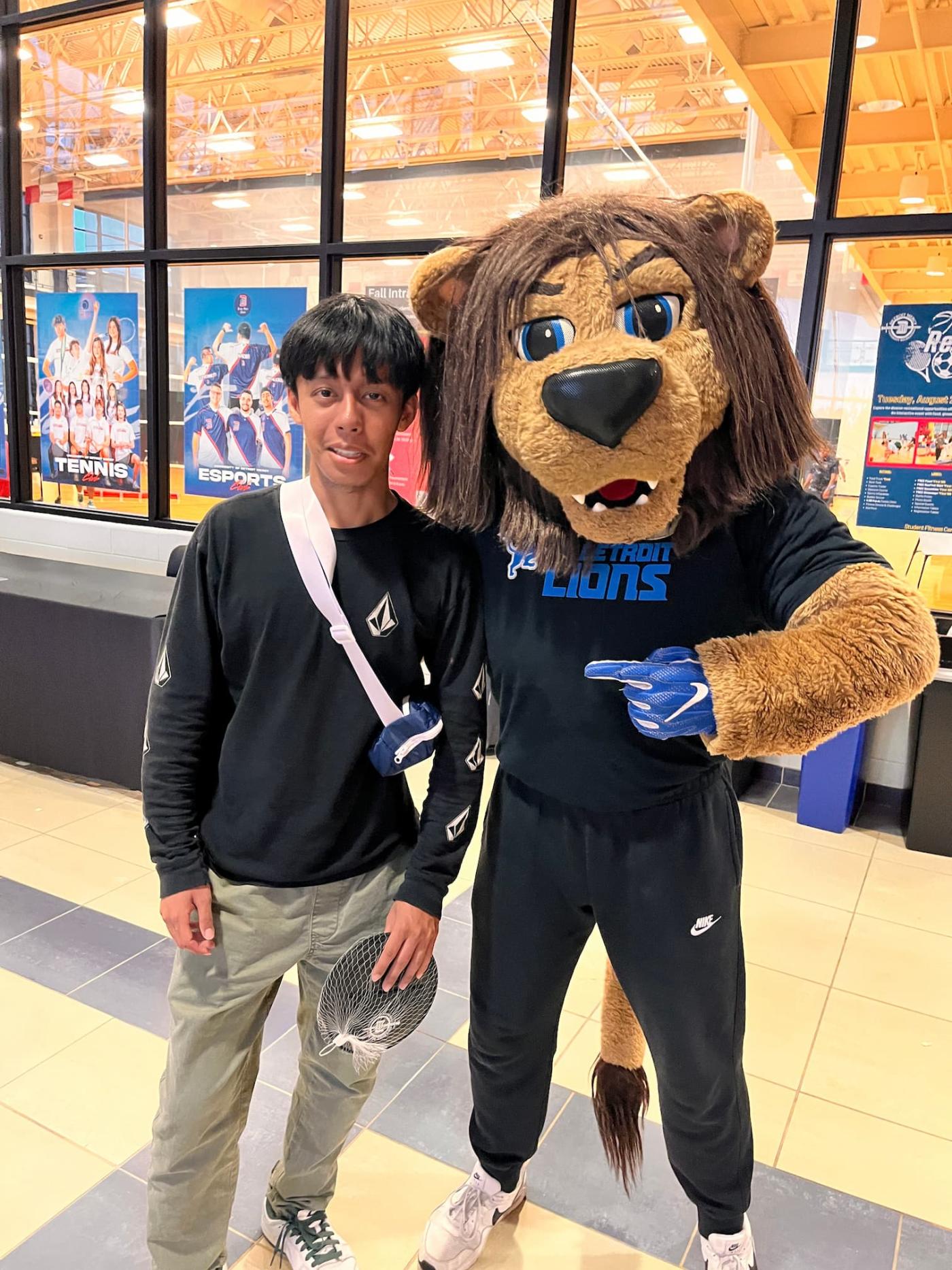 student stands next to a Detroit Lions mascot in a furry costume 