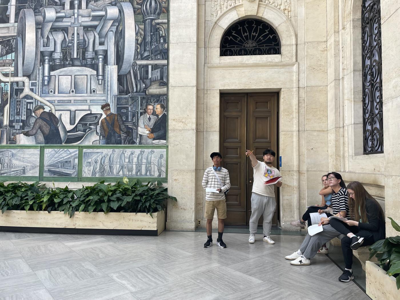 A student points across a mural room in the Detroit Institute of Arts while other students look on 