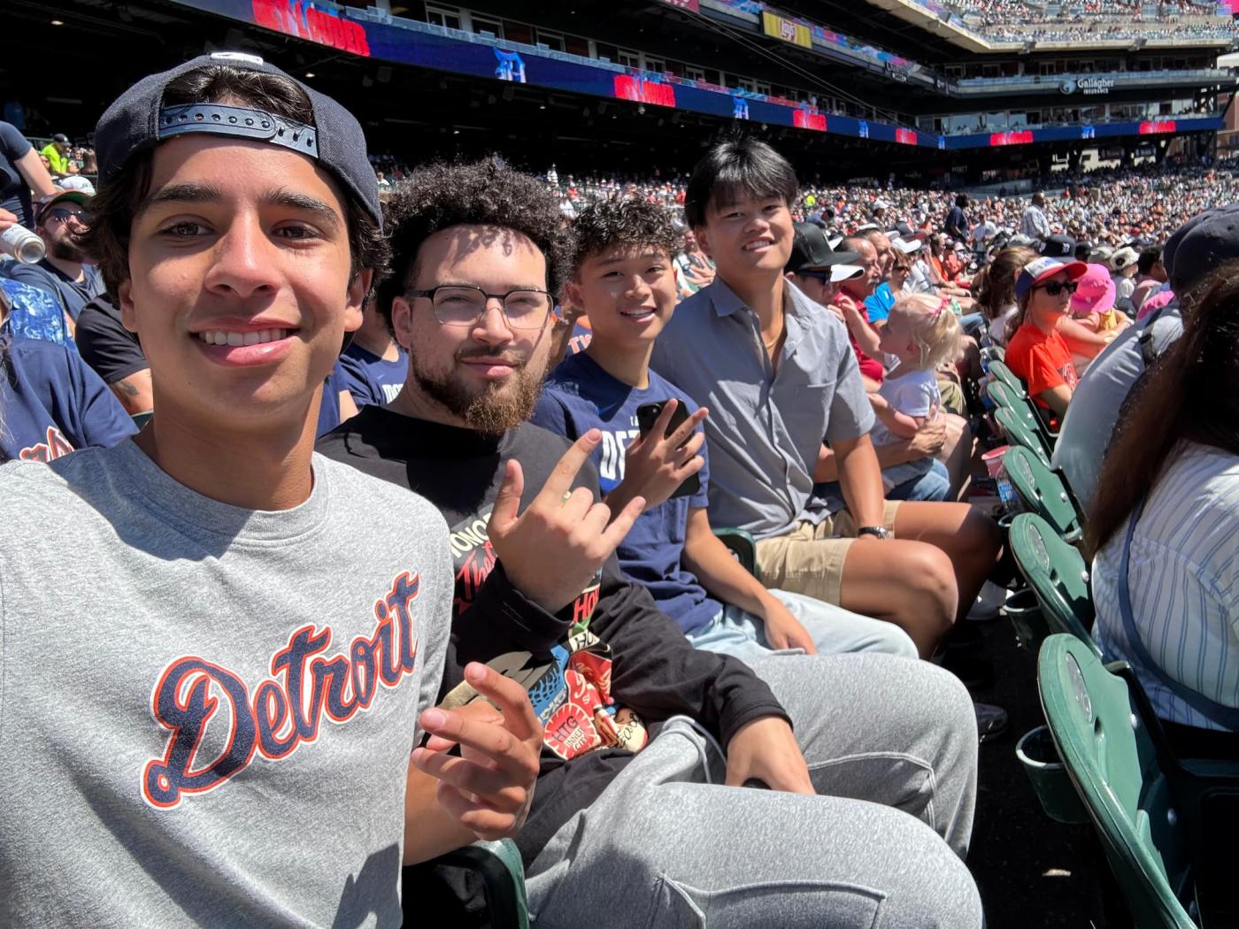 Four male students look toward the camera in the stands of a Detroit Tigers baseball game. 