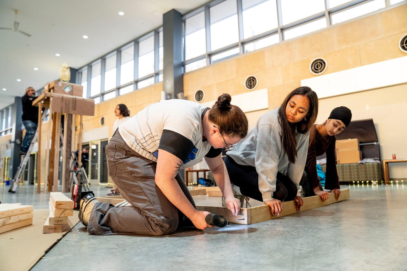 Two students hold a 2x4 wood plank in place while a third student screws another plank into the side at a 90 degree angle
