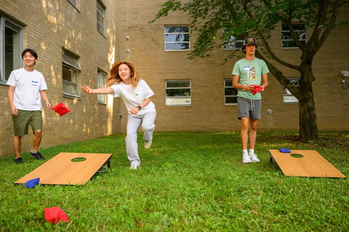 A woman throws a bean bag in a game of bean bag toss. Two men stand on either side of her. 