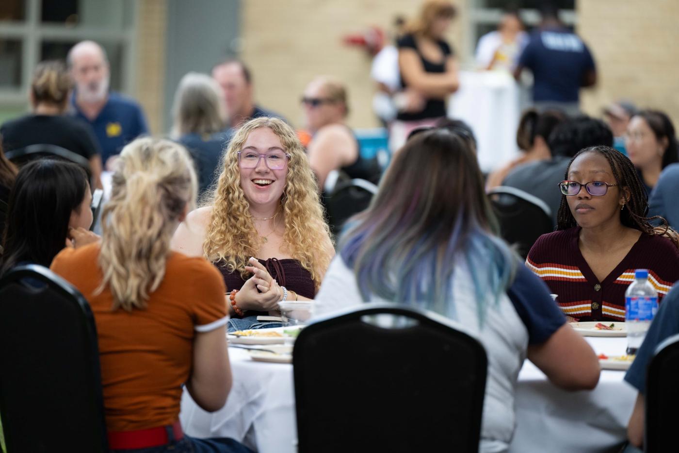 Five students sit around a dinner table in a courtyard talking and smiling. 