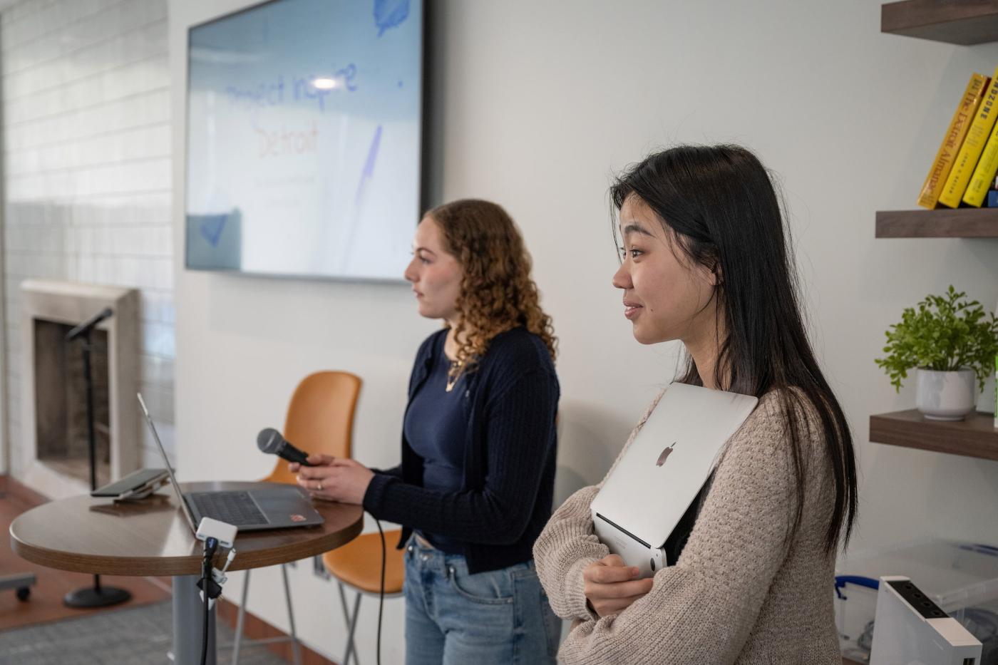 Two students present at the front of a classroom, one holding a laptop, the other holding a microphone