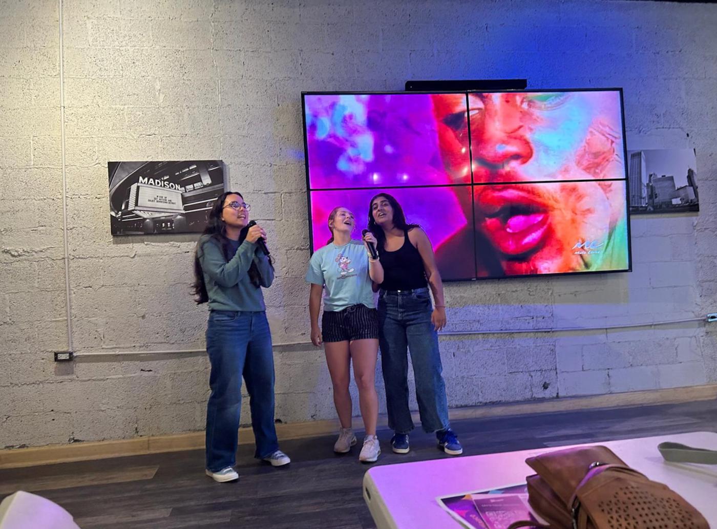 Three women sing into microphones with a large colorful screen behind them. 