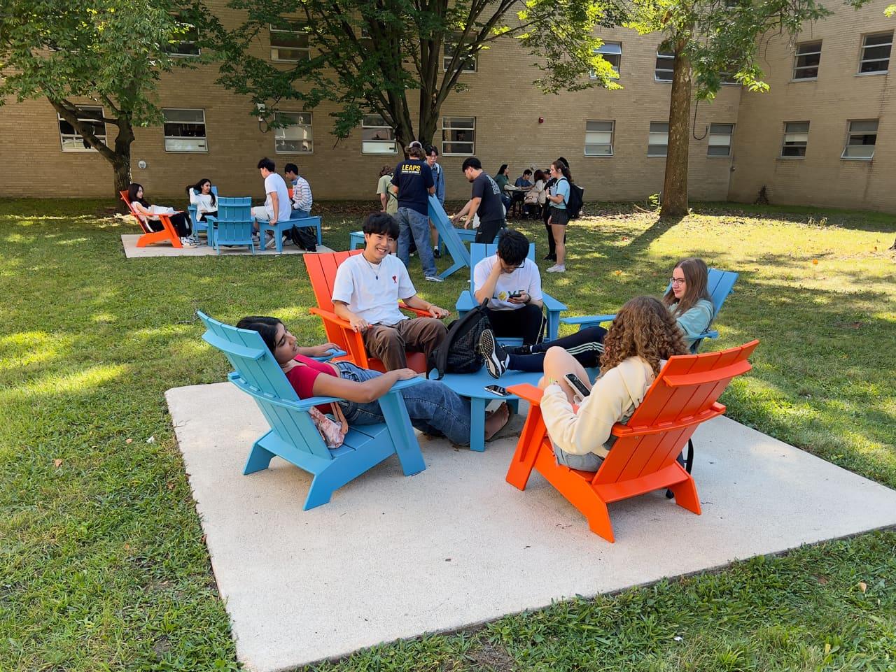 In the foreground, five students recline in colorful chairs outdoors. In the background, other students are gathering. 