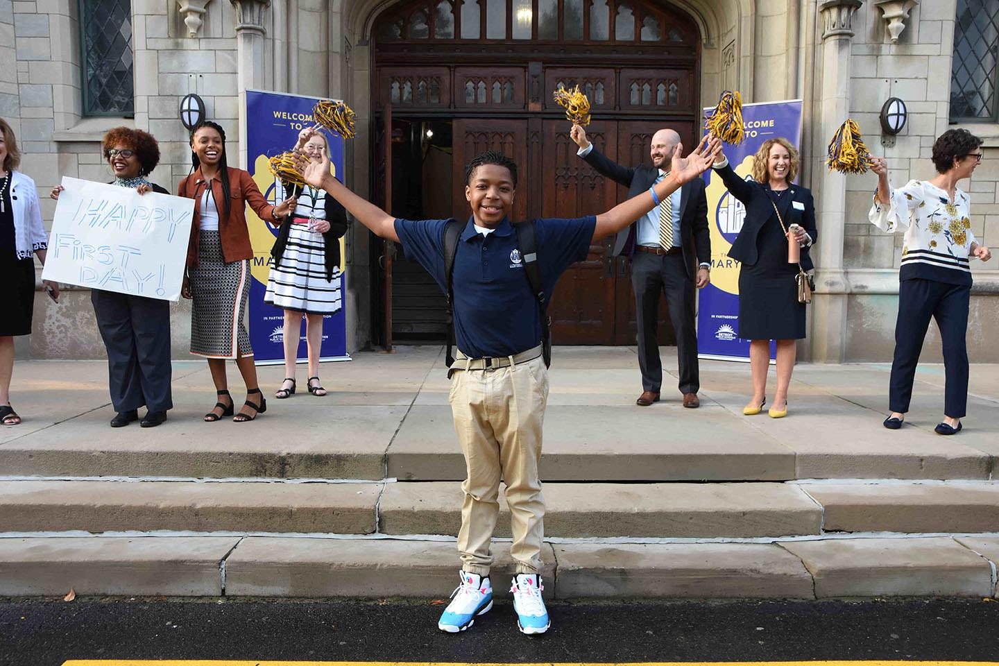 A high school freshman stands in front of his new school with his arms outstretched. School administrators and P-20 partnership staff stand behind him, cheering him on.