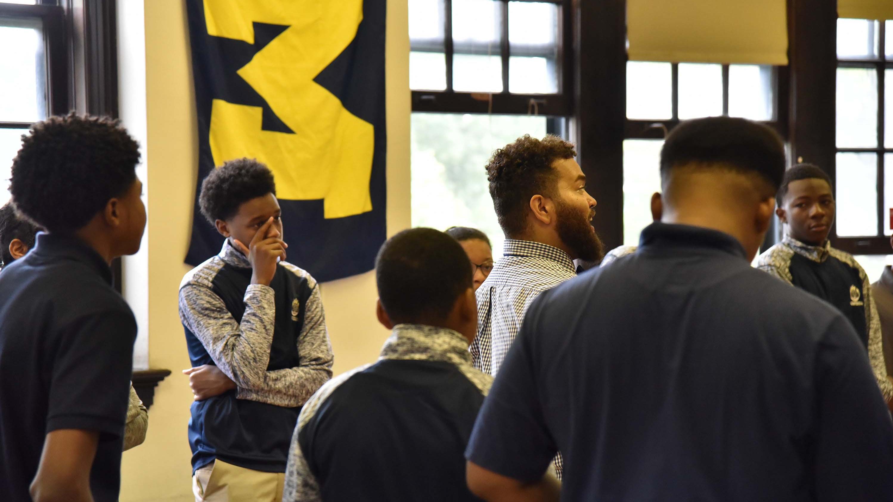 A teacher surrounded by students in a room with a large University of Michigan flag on the wall