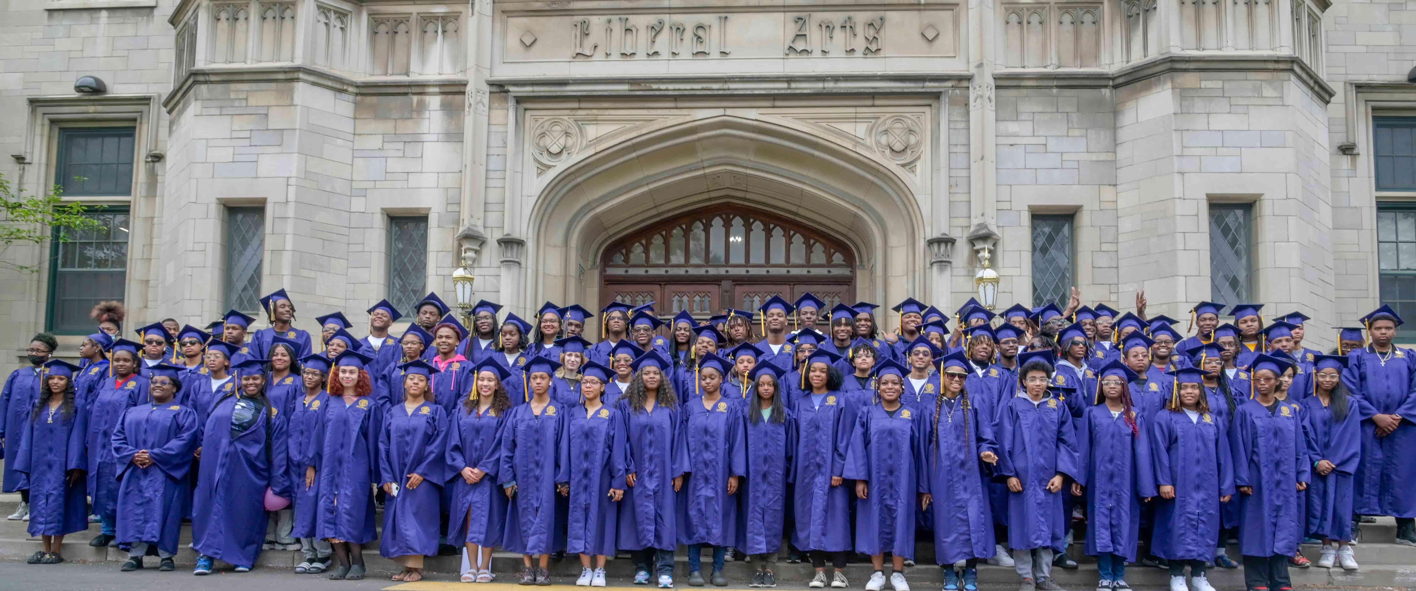 Approximately 100 high school seniors wearing blue graduation gowns stand outside of their high school.