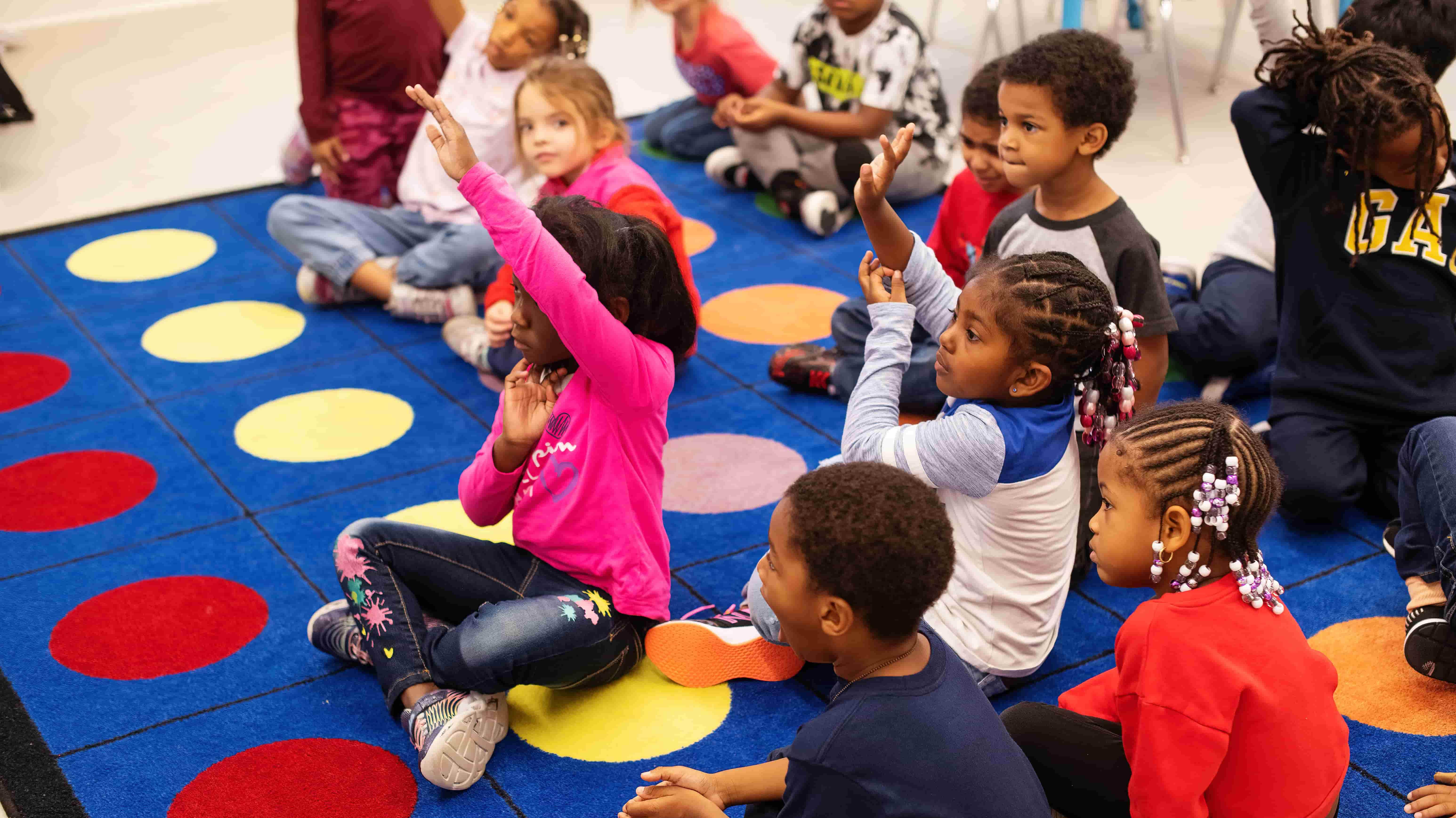 Approximately a dozen young children sit on a colorful rug. Some of the children are raising their hands.