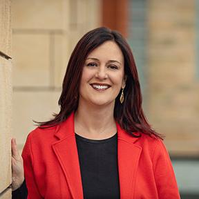 Professional headshot of Dr. Rebecca Quintana, who is wearing a red blazer and leaning against a brick wall.