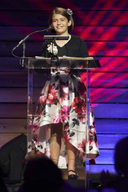 Olive Martin (Magazine, Fall 2021) Photo of Olive Martin. Olive wears a dress with a black top and floral bottom, and smiles while standing in front of a podium.