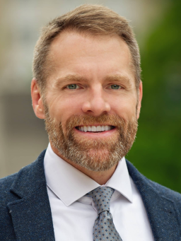 Headshot of adult wearing a suit jacket and tie, smiling warmly.