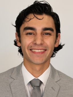 Headshot of adult in a sand-colored suit and tie, smiling.