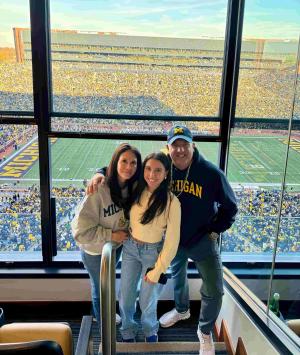 Two parents stand on either side of their college-age daughter with their arms around each other. Through the windows behind them is a view into the Michigan stadium, packed with people for a football game.