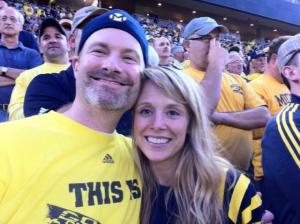 A smiling man with a beard wearing a yellow t-shirt and a blue ski cap poses with his arm around a smiling woman with long blond hair. Behind them rows of spectators wear maize and blue sports attire.
