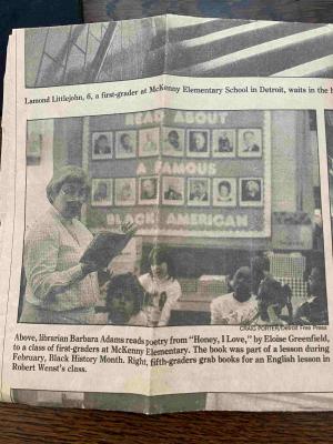 A black and white clipping from an old issue of the Detroit Free Press shows a teacher wearing glasses and reading from an open book to a roomful of students. Behind them is a display board with portraits and letters that read “Read about a famous Black American.”
