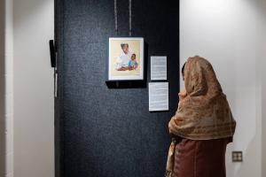 A visitor to a gallery wears a headscarf and looks at a framed portrait on the wall. The portrait is of an older person holding a young child on their lap.