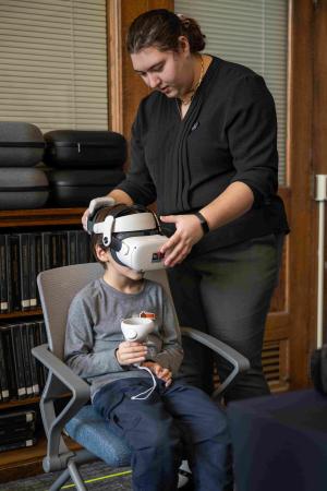 A woman in a black blouse stands, adjusting the VR headset on an elementary school-aged child who sits next to her in a chair. The child clutches a console connected to the headset, and appears excited to try out the futuristic equipment. 