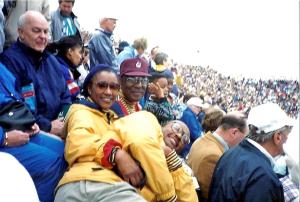 Seated in a sports stadium, a middle aged couple wearing maize and blue sports attire smile at the camera as a grinning young man stretches across their lap with his arms crossed. The woman has her arm around his waist. 